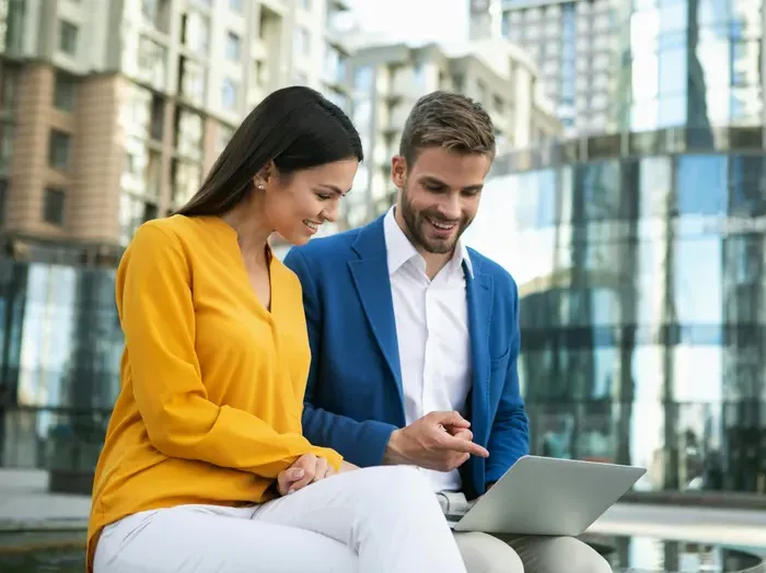 Two professionals meeting outdoors by offices, smiling while reviewing a proposal on a laptop and planning work